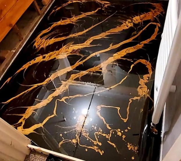 The image depicts a stunning floor finished with black matte epoxy resin, adorned with intricate golden metallic pigment swirls. The contrast between the deep black and shimmering gold creates a luxurious and dynamic appearance
